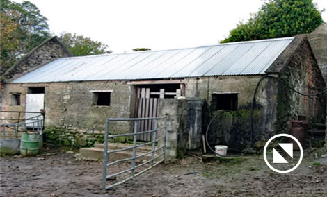 Old stone barn with a metal roof, surrounded by a muddy yard and a metal gate in the foreground.
